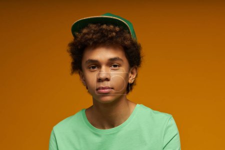 Téléchargez les photos : Adolescent mignon avec une expression faciale neutre portant un t-shirt vert et un bonnet regardant la caméra tout en posant sur fond jaune en isolement - en image libre de droit