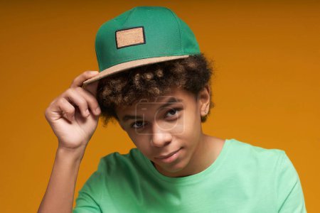 Foto de Tranquilo adolescente en camiseta tocando visor de gorra de béisbol verde y mirando a la cámara mientras posa durante la sesión de fotos en aislamiento - Imagen libre de derechos