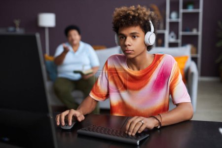 Photo for Serious diligent teenager in headphones clicking mouse and typing on computer keyboard while sitting by desk and doing homework - Royalty Free Image