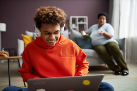 Téléchargez les photos : Adolescent joyeux mignon avec ordinateur portable préparer la tâche à la maison et rire tout en écoutant des choses drôles dans les écouteurs - en image libre de droit