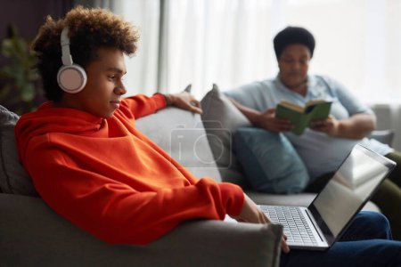 Téléchargez les photos : Vue latérale d'un adolescent dans un casque et un sweat à capuche rouge regardant un film ou une vidéo en ligne sur un écran d'ordinateur portable contre un livre de lecture de femme - en image libre de droit