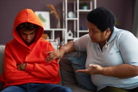 Téléchargez les photos : Femme mûre afro-américaine parlant à un petit-fils adolescent boudé ou stressé en sweat à capuche rouge assis sur le canapé à côté de lui - en image libre de droit
