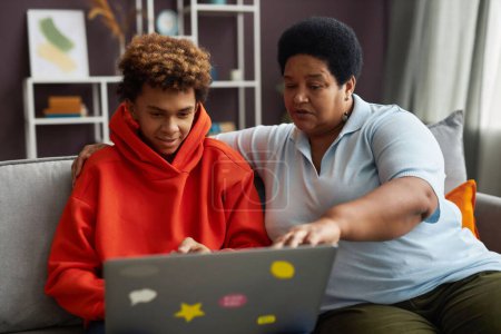 Foto de Mujer madura afroamericana y adolescente diligente mirando la pantalla del ordenador portátil mientras ve video en línea o discute información - Imagen libre de derechos