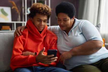 Téléchargez les photos : Afro-américain adolescent garçon en rouge sweat à capuche montrant vidéo en ligne dans smartphone à sa grand-mère tandis que les deux assis sur canapé - en image libre de droit