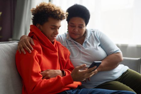 Foto de Feliz adolescente con capucha roja y su madre mirando la pantalla del teléfono inteligente mientras ve y discute el video en línea en el ocio - Imagen libre de derechos