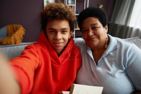 Téléchargez les photos : Femme mûre affectueuse et son fils adolescent prenant selfie à la maison tout en étant assis sur le canapé dans le salon et en regardant la caméra - en image libre de droit