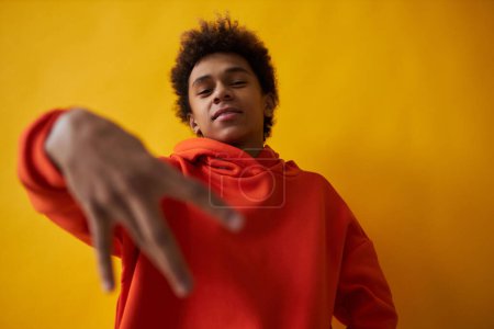 Téléchargez les photos : Jeune Afro-Américain en sweat à capuche rouge regardant la caméra tout en se tenant sur fond jaune et en gardant la main droite devant lui - en image libre de droit
