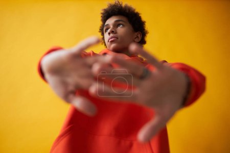 Téléchargez les photos : Mignon afro-américain adolescent garçon en sweat à capuche rouge étirant les bras en face de lui tout en posant sur fond jaune lors de la séance photo - en image libre de droit
