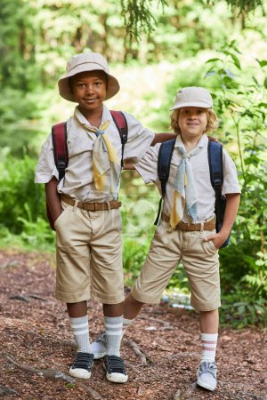 Téléchargez les photos : Portrait vertical pleine longueur de deux scouts garçons mignons regardant la caméra à l'extérieur en forêt tout en profitant d'une sortie éducative - en image libre de droit