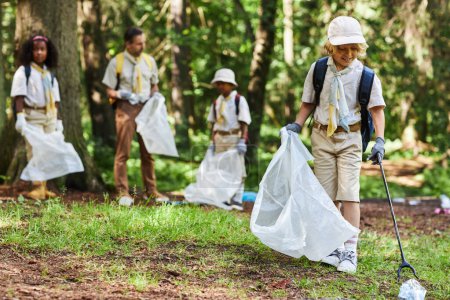 Foto de Retrato de cuerpo entero del lindo boy scout ayudando a limpiar el bosque y sosteniendo la bolsa de basura, espacio para copiar - Imagen libre de derechos