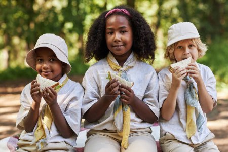 Téléchargez les photos : Vue de face au groupe de scouts profitant de la pause déjeuner pendant le voyage de randonnée avec un accent sur la fille noire souriant - en image libre de droit