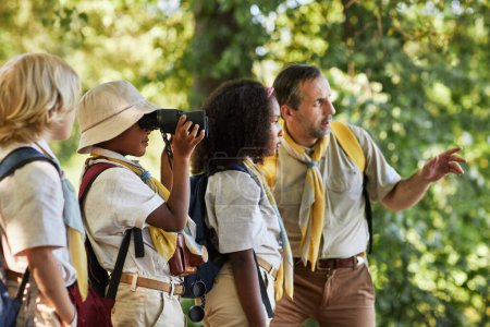 Foto de Vista lateral al diverso grupo de exploradores en el bosque explorando la naturaleza y mirando en prismáticos - Imagen libre de derechos