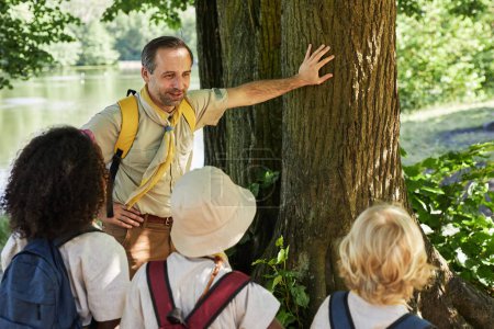 Téléchargez les photos : Portrait en grand angle d'un homme adulte souriant en tant que chef scout parlant aux enfants dans la forêt et s'appuyant sur un arbre - en image libre de droit