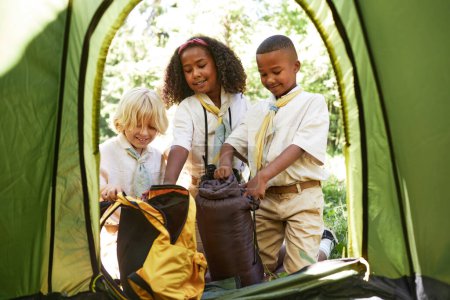 Photo for Diverse group of young scouts putting up tent in forest, framed by tent door - Royalty Free Image