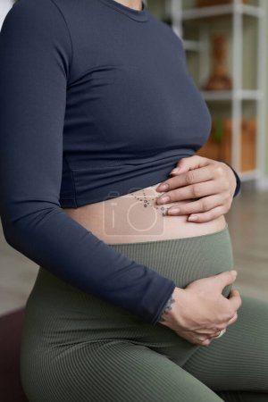 Foto de Primer plano vertical de la mujer embarazada haciendo yoga prenatal con enfoque en las manos que sostienen el vientre tatuado - Imagen libre de derechos