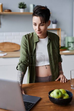 Photo for Vertical portrait of pregnant young woman with tattooed using laptop on kitchen counter - Royalty Free Image