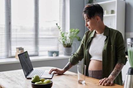 Photo for Side view portrait of pregnant young woman using laptop standing in cozy kitchen and checking work messages - Royalty Free Image