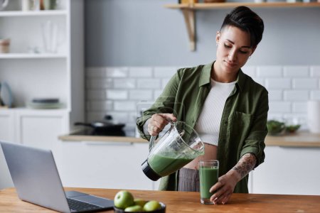Foto de Retrato de la cintura hacia arriba de la joven embarazada haciendo batido verde para el desayuno en un entorno de cocina mínima - Imagen libre de derechos