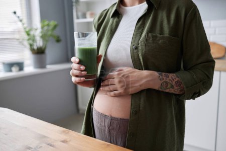 Téléchargez les photos : Gros plan de la jeune femme enceinte buvant smoothie vert le matin en mettant l'accent sur le ventre, espace de copie - en image libre de droit