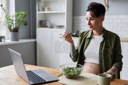 Téléchargez les photos : Portrait de la femme enceinte moderne manger de la salade dans la cuisine et regarder la vidéo éducative en ligne - en image libre de droit
