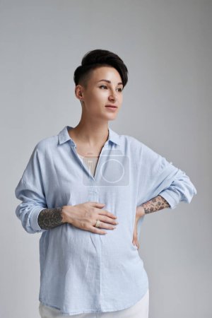 Téléchargez les photos : Portrait minime de jeune femme enceinte tatouée posant sur fond gris et détournant les yeux - en image libre de droit