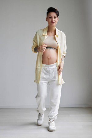 Téléchargez les photos : Portrait complet de jeune femme enceinte portant des vêtements décontractés confortables en studio minimal et regardant la caméra - en image libre de droit