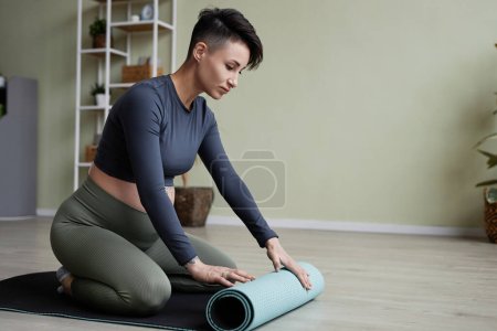 Téléchargez les photos : Portrait minimal de la jeune femme enceinte portant une tenue de sport se préparant pour le cours de yoga prénatal et déroulant tapis - en image libre de droit