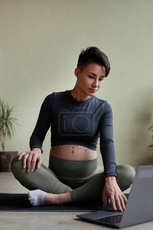 Foto de Retrato vertical de una mujer joven embarazada usando un portátil listo para el entrenamiento prenatal en línea de yoga en casa - Imagen libre de derechos