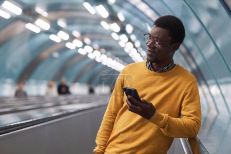 Téléchargez les photos : Jeune homme d'affaires afro-américain utilisant un smartphone tout en restant debout sur l'escalator dans le métro - en image libre de droit