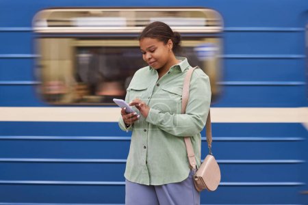 Téléchargez les photos : Femme afro-américaine tapant message sur smartphone tout en se tenant dans le métro avec le train en arrière-plan - en image libre de droit