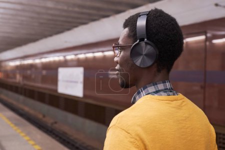 Téléchargez les photos : Vue arrière de l'homme afro-américain dans des écouteurs sans fil écoutant de la musique debout dans la station dans le métro - en image libre de droit