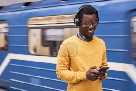 Foto de Sonriente hombre en auriculares inalámbricos escribiendo mensaje en el teléfono móvil de pie en la estación con el tren en segundo plano - Imagen libre de derechos