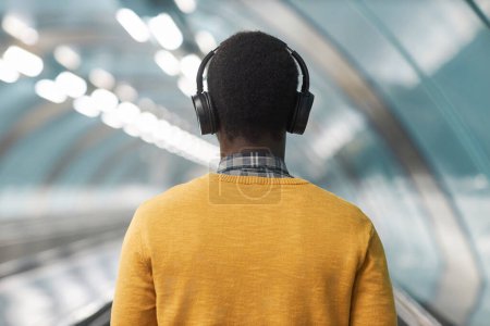 Foto de Vista trasera del hombre afroamericano con auriculares inalámbricos escuchando música mientras está de pie en la escalera mecánica en el metro - Imagen libre de derechos