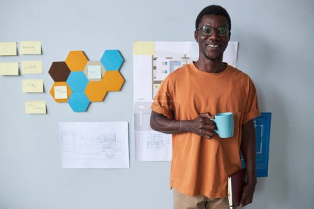 Foto de Retrato del joven arquitecto afroamericano sosteniendo una taza de café y sonriendo a la cámara mientras está de pie en la oficina - Imagen libre de derechos