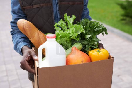 Téléchargez les photos : Gros plan de jeune homme noir portant boîte avec des légumes frais tout en livrant des produits d'épicerie à la porte - en image libre de droit