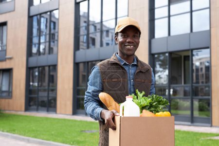 Téléchargez les photos : Portrait taille haute de jeune homme noir portant une boîte avec des légumes frais tout en livrant des produits d'épicerie à la porte et en souriant à la caméra, en copiant l'espace - en image libre de droit