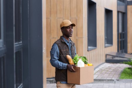 Foto de Retrato de cintura hacia arriba del joven negro que lleva la caja con comestibles al aire libre mientras trabaja en la entrega de alimentos - Imagen libre de derechos