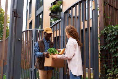 Téléchargez les photos : Portrait d'un jeune homme noir distribuant une boîte avec des produits alimentaires à un client à l'extérieur à la porte d'un complexe d'appartements - en image libre de droit