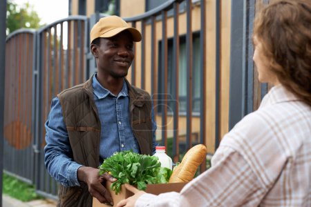 Foto de Retrato de cintura hacia arriba de un joven negro entregando una caja con alimentos frescos a una mujer joven en la ciudad - Imagen libre de derechos