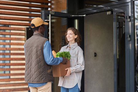 Photo for Waist up portrait of smiling young woman holding box with groceries outdoors and talking to delivery man - Royalty Free Image