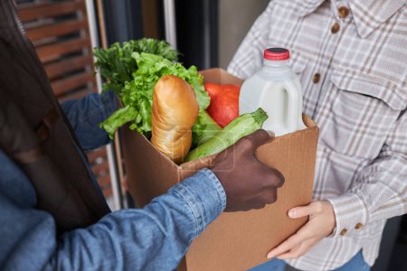Photo for Closeup of young woman holding box with groceries outdoors with delivery man - Royalty Free Image