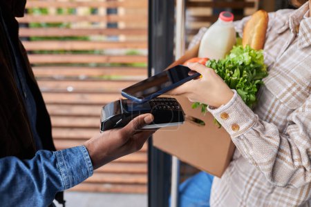 Téléchargez les photos : Gros plan d'une femme méconnaissable payant pour la livraison d'épicerie via smartphone à l'extérieur - en image libre de droit