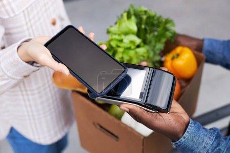 Téléchargez les photos : Gros plan de la jeune femme payant pour la livraison d'épicerie via smartphone NFC - en image libre de droit