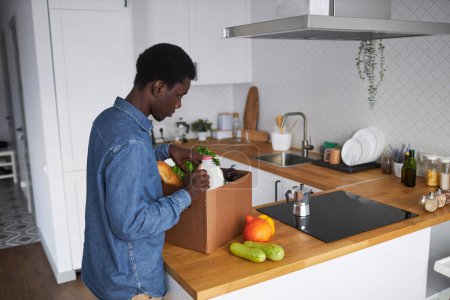 Foto de Retrato de vista lateral del joven negro desempacando comestibles frescos en el interior de la cocina, espacio para copiar - Imagen libre de derechos