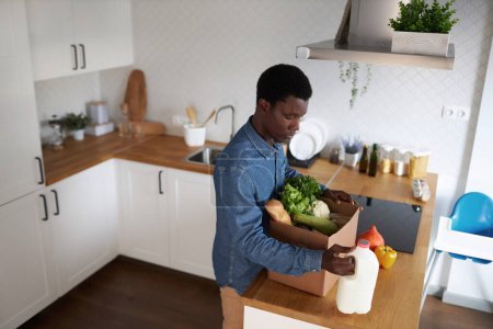 Foto de Vista de ángulo alto en el joven hombre negro desembalaje de comestibles frescos en el interior de la cocina, espacio de copia - Imagen libre de derechos