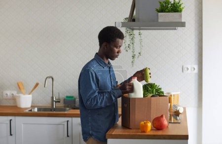 Foto de Retrato de vista lateral del hombre negro desempacando comestibles frescos en el interior de la cocina mínima, espacio de copia - Imagen libre de derechos