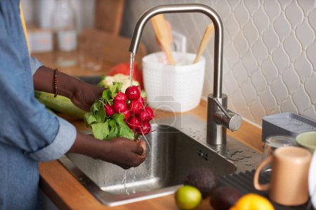 Photo for Side view closeup of black man washing fresh radishes and vegetables in tap water, copy space - Royalty Free Image
