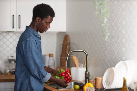 Foto de Retrato de vista lateral del joven negro lavando verduras frescas en la cocina, espacio para copiar - Imagen libre de derechos
