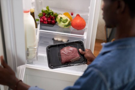 Téléchargez les photos : Gros plan de jeune homme noir sortant le bœuf du réfrigérateur pendant la cuisson du dîner à la maison - en image libre de droit