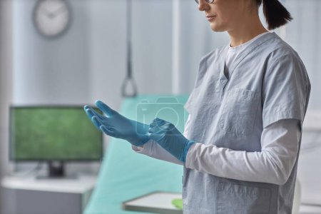 Téléchargez les photos : Jeune infirmière en uniforme portant des gants de protection avant le travail debout dans la salle d'hôpital - en image libre de droit